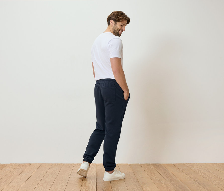 Férfi fehér pólóban és kék tréningnadrágban áll egy fapadlón.