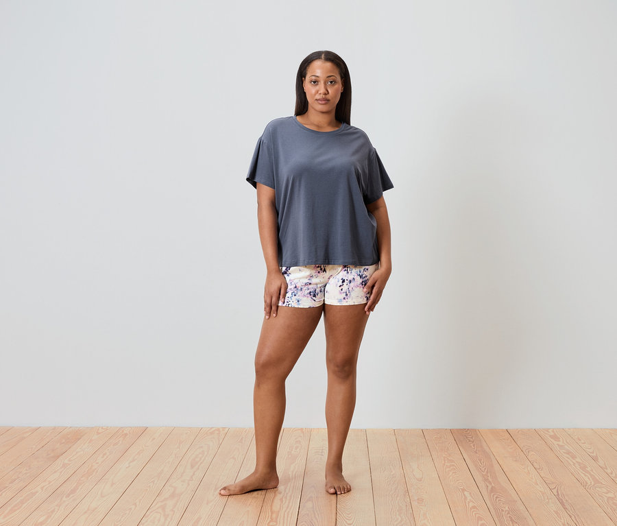 Egy nő mezítláb áll egy fapadlón, szürke pólót és virágmintás rövidnadrágot visel.