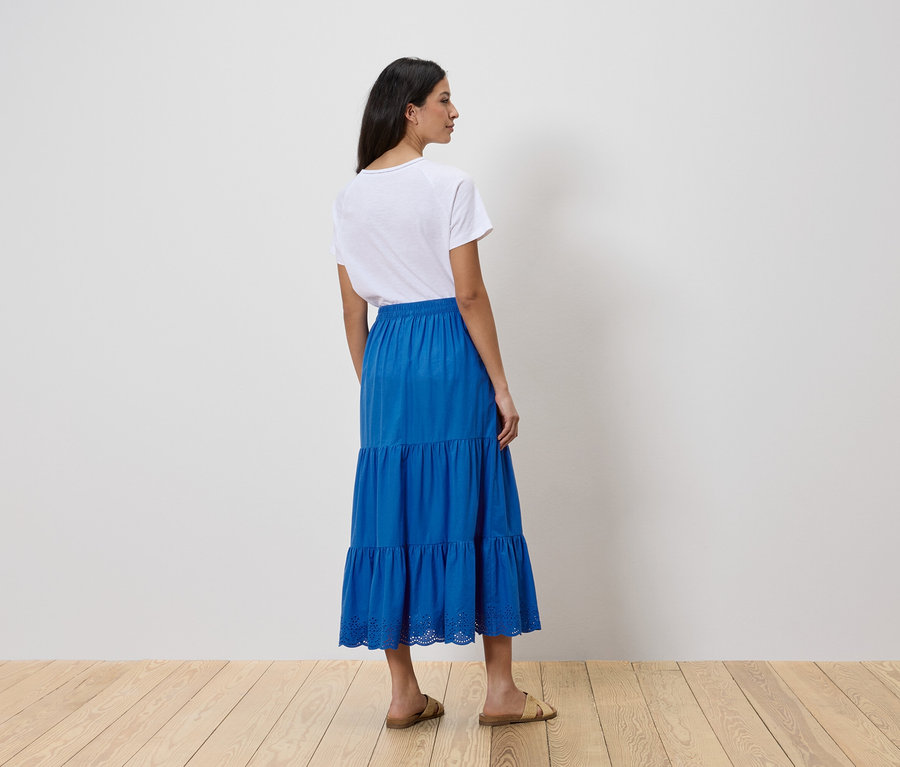 Egy nő háttal áll a kamerának, fehér pólót és kék, réteges szoknyát visel.