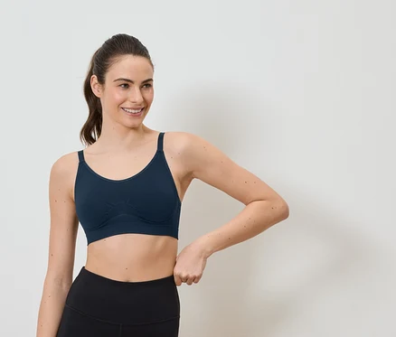 Mosolygó, lófarokba kötött hajú nő sötétkék sportmelltartót és fekete leggingset visel fehér háttér előtt.