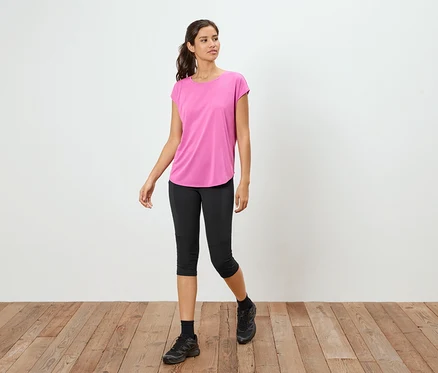 Nő áll a fapadlón, rózsaszín pólót, fekete capri leggingset és fekete tornacipőt visel.
