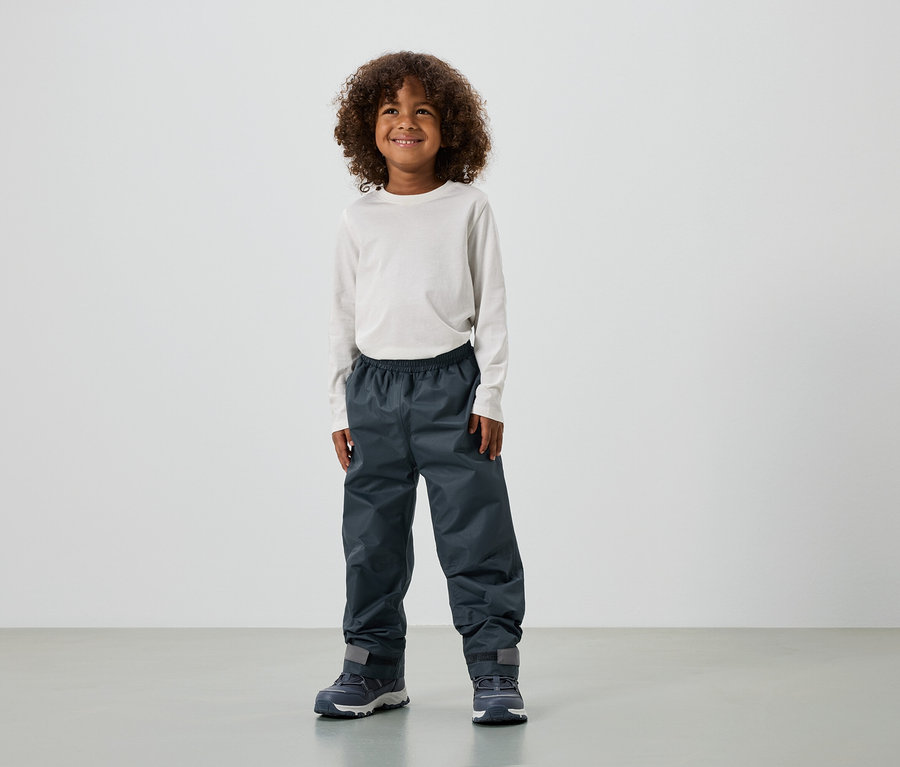 Egy mosolygó, göndör hajú fiú hosszú ujjú felsőt, esőnadrágot és edzőcipőt visel.