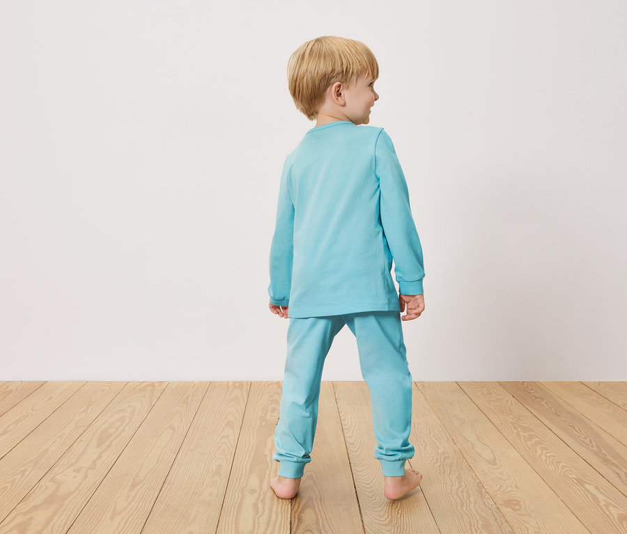 Kisfiú hátulról, világoskék pizsamában, fa padlón áll egy fehér fal előtt.