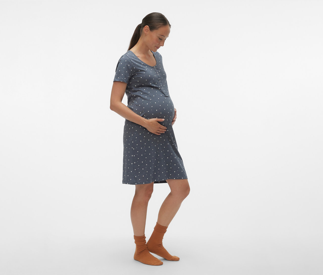 Profilból látható terhes nő kék, fehér pöttyös MAMALICIOUS kismama- és szoptatós hálóinget és barna zoknit visel.