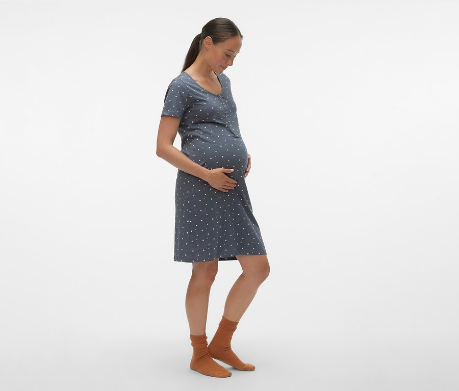 Profilból látható terhes nő kék, fehér pöttyös MAMALICIOUS kismama- és szoptatós hálóinget és barna zoknit visel.
