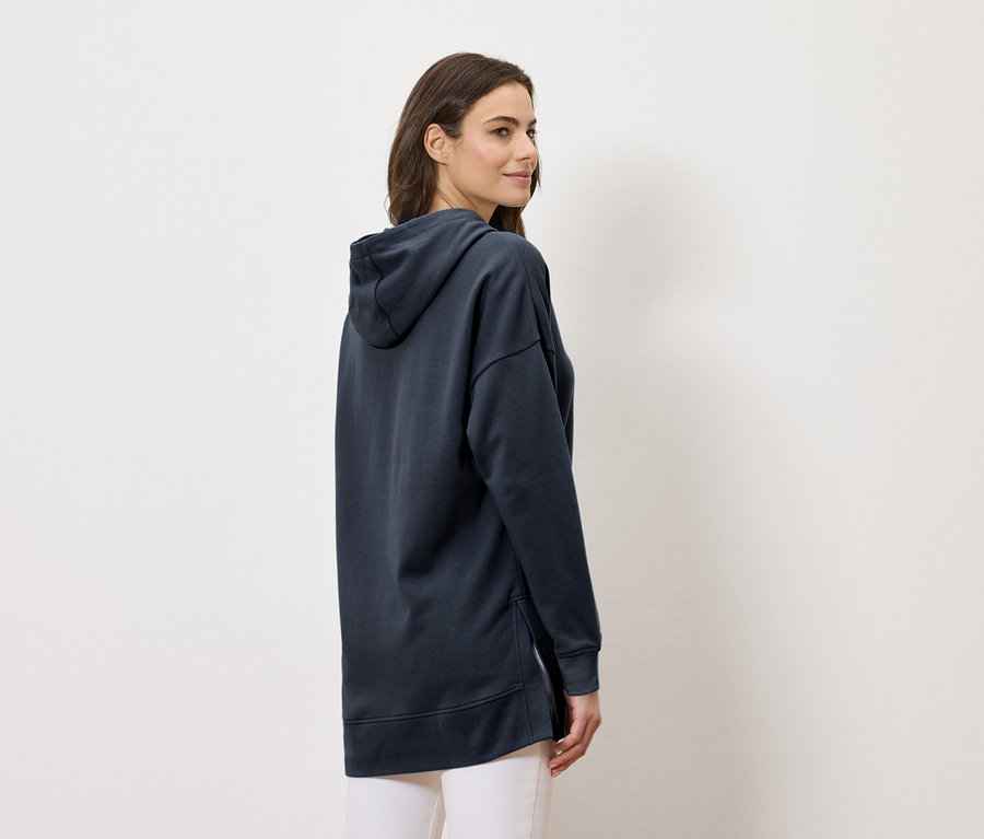 Nő visel egy hosszú, sötétkék kapucnis pulóvert és fehér nadrágot, oldalnézetből.