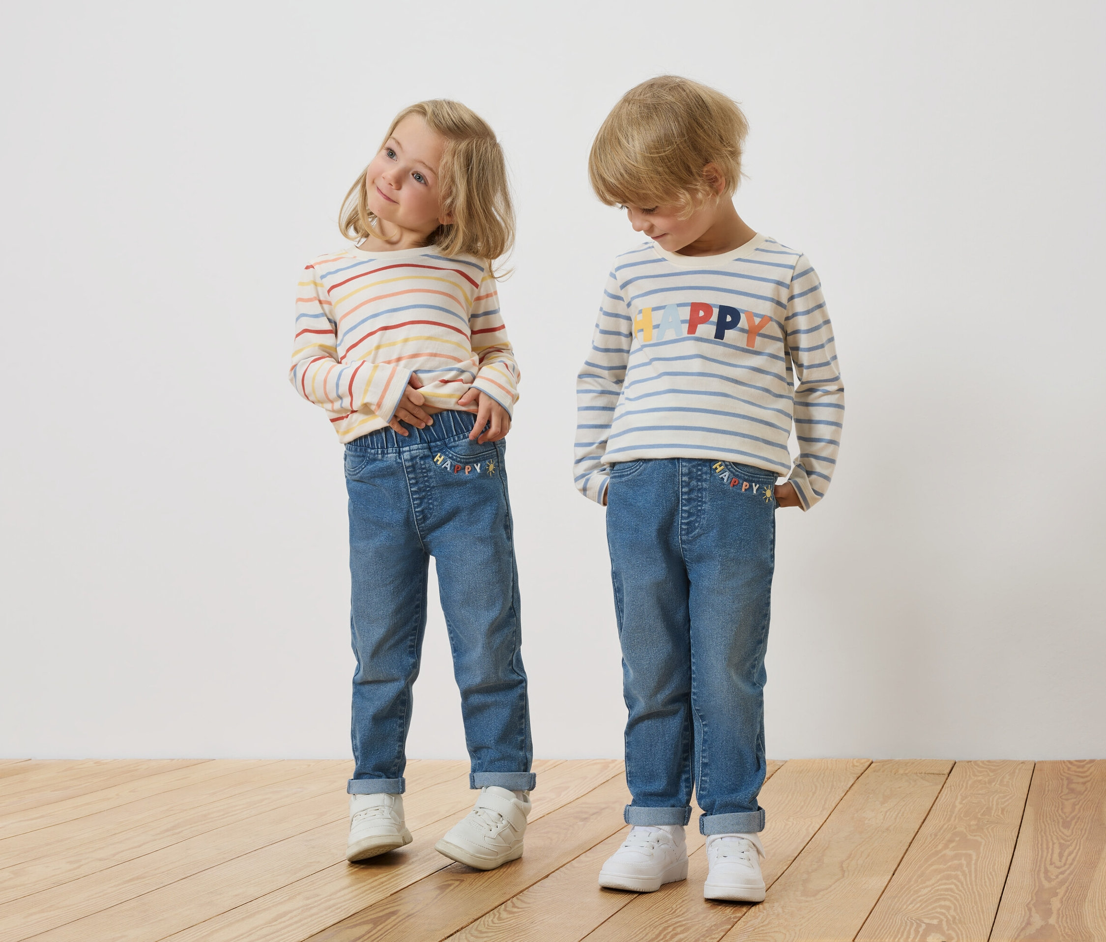 Egy szőke kislány és egy szőke kisfiú áll egymás mellett, csíkos, hosszú ujjú felsőket és farmert viselnek.