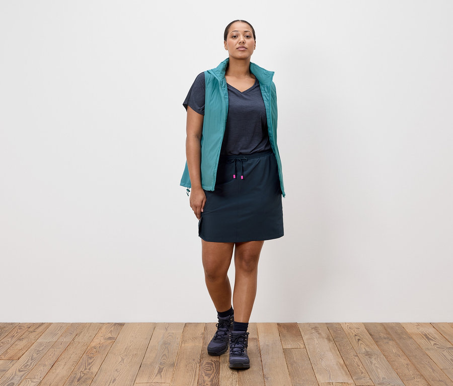 Egy nő áll egy fehér fal előtt, tengerészkék szoknyát, tengerészkék pólót, petrolkék mellényt és tengerészkék túrabakancsot visel.