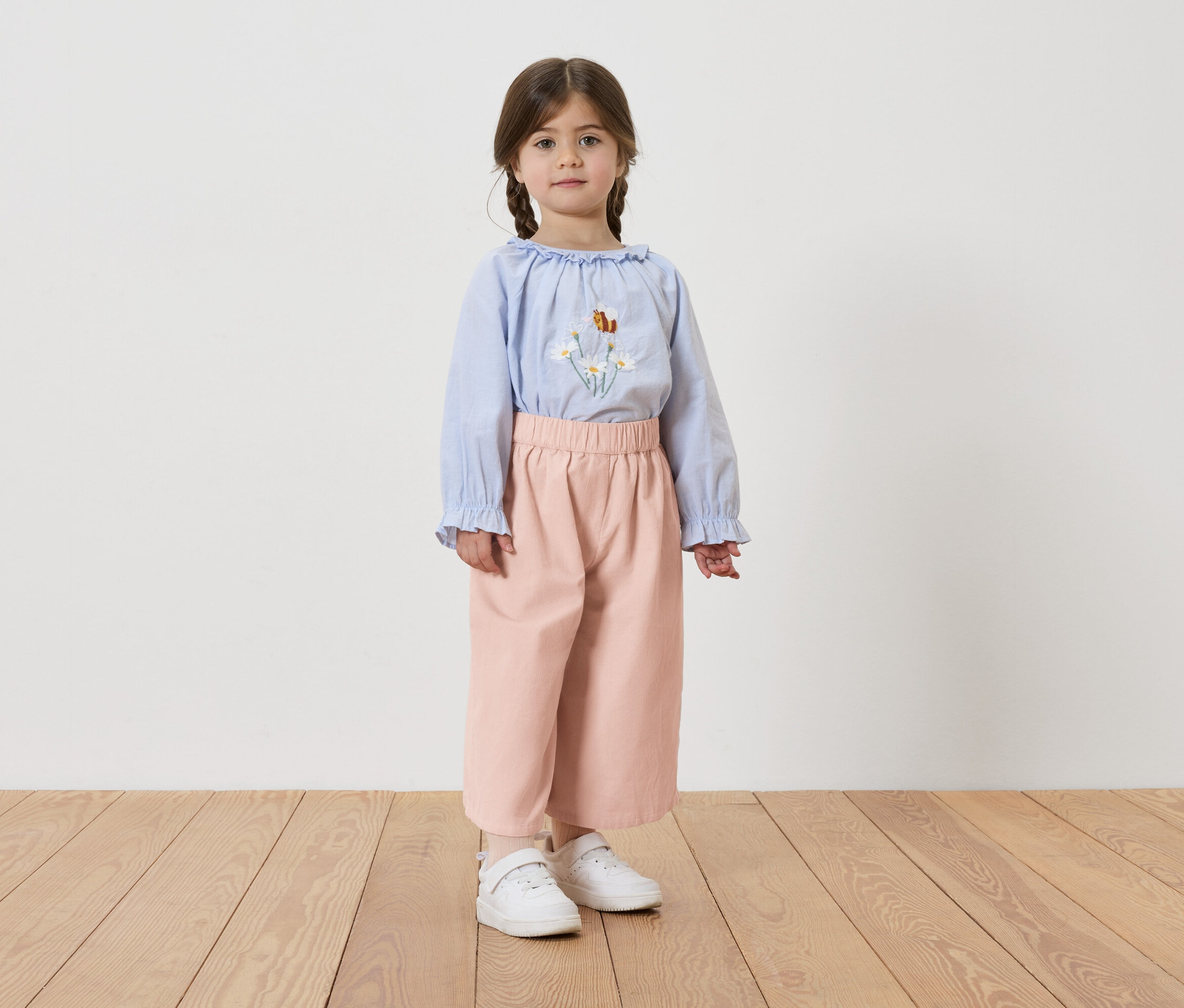 Lány copfokkal, világoskék, virágmintás blúzt, rózsaszín nadrágot és fehér tornacipőt visel.