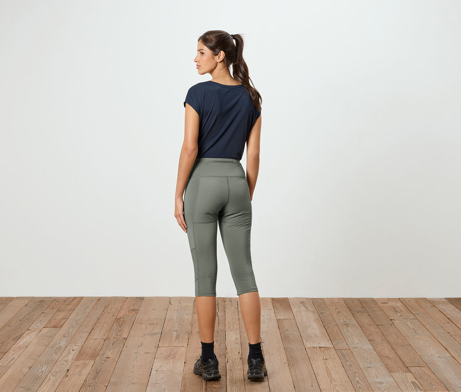 Hátulról látható lófarokos nő tengerészkék pólót és olívazöld capri leggingset visel egy fehér fal előtt.