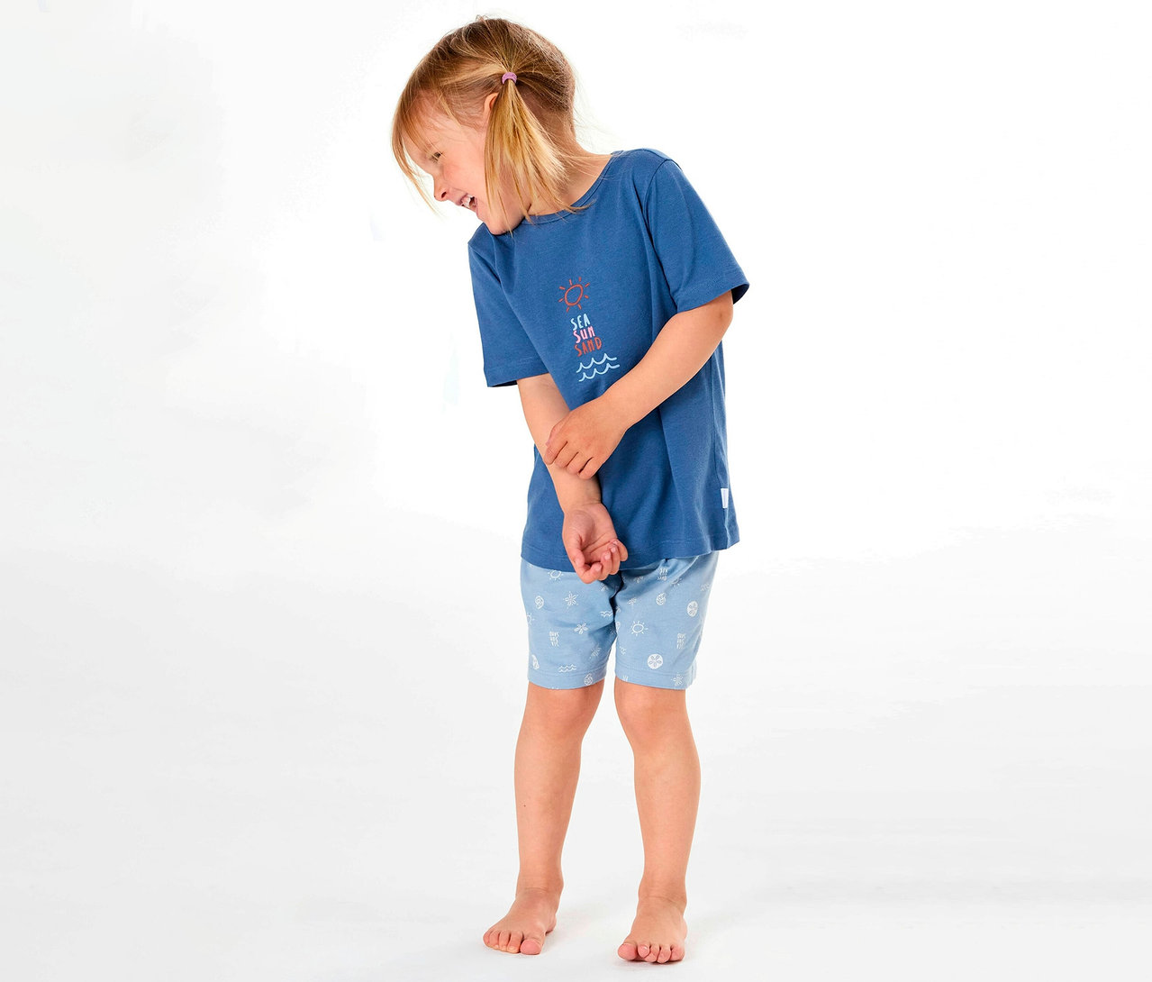 Kislány SCHIESSER rövidnadrágos gyerekpizsamát visel kék pólóval és mintás rövidnadrággal, oldalra mosolyogva.