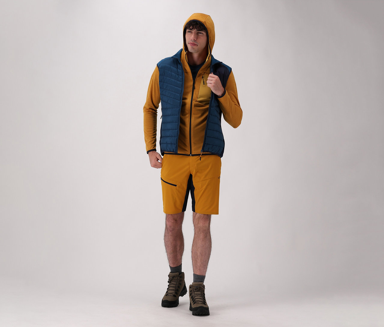 Férfi visel CMP férfi kapucnis felsőt, CMP hibrid férfimellényt, rövidnadrágot és csizmát világos háttér előtt.