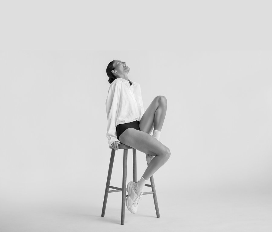 Egy nő mosolyogva ül egy sámlin, fehér Hosszú lenvászon ingblúz, fekete alsónemű, fehér zokni és fehér tornacipő van rajta.