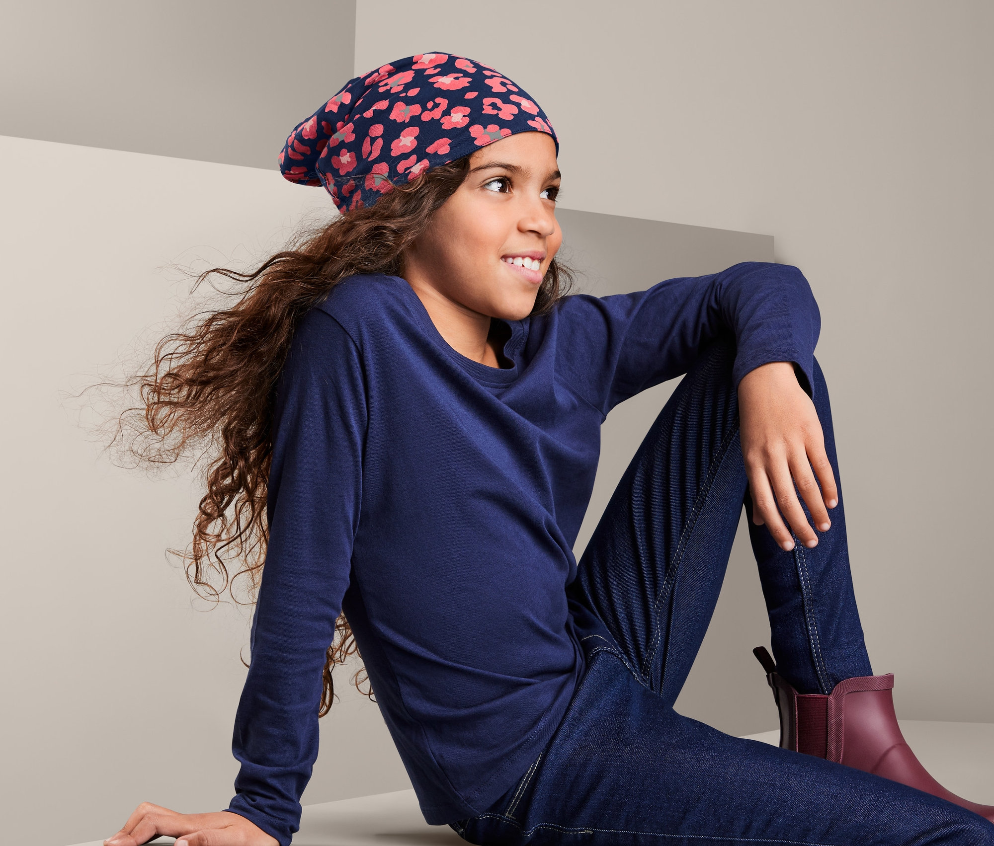 Egy lány ül, és virágmintás kifordítható sapkát, kék hosszú ujjú pólót, farmert és bordó csizmát visel.