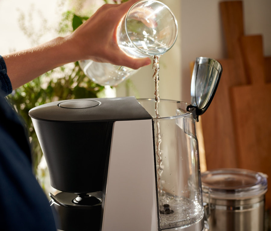Egy nő vizet önt egy átlátszó pohárból egy szürke-fekete »Let's Brew« filteres kávéfőzőbe.