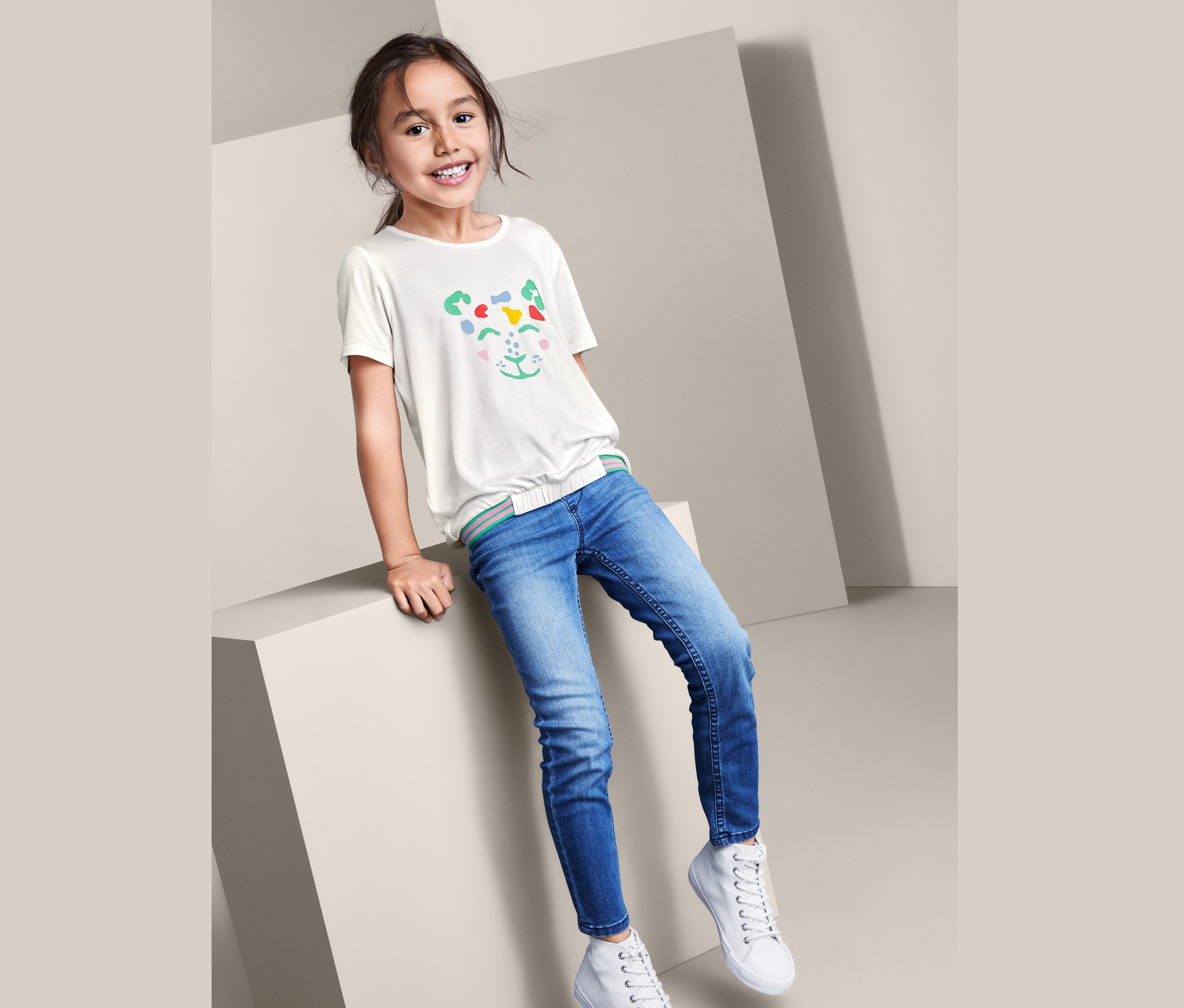 Egy lány ül egy kockán, farmerben, fehér tornacipőben és egy állatarcú, kislány pólóban.