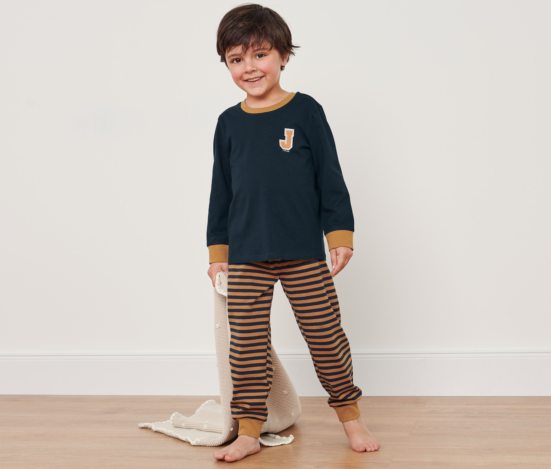 Fiúgyermek kék-barna pizsamában áll a padlón és egy takarót tart.