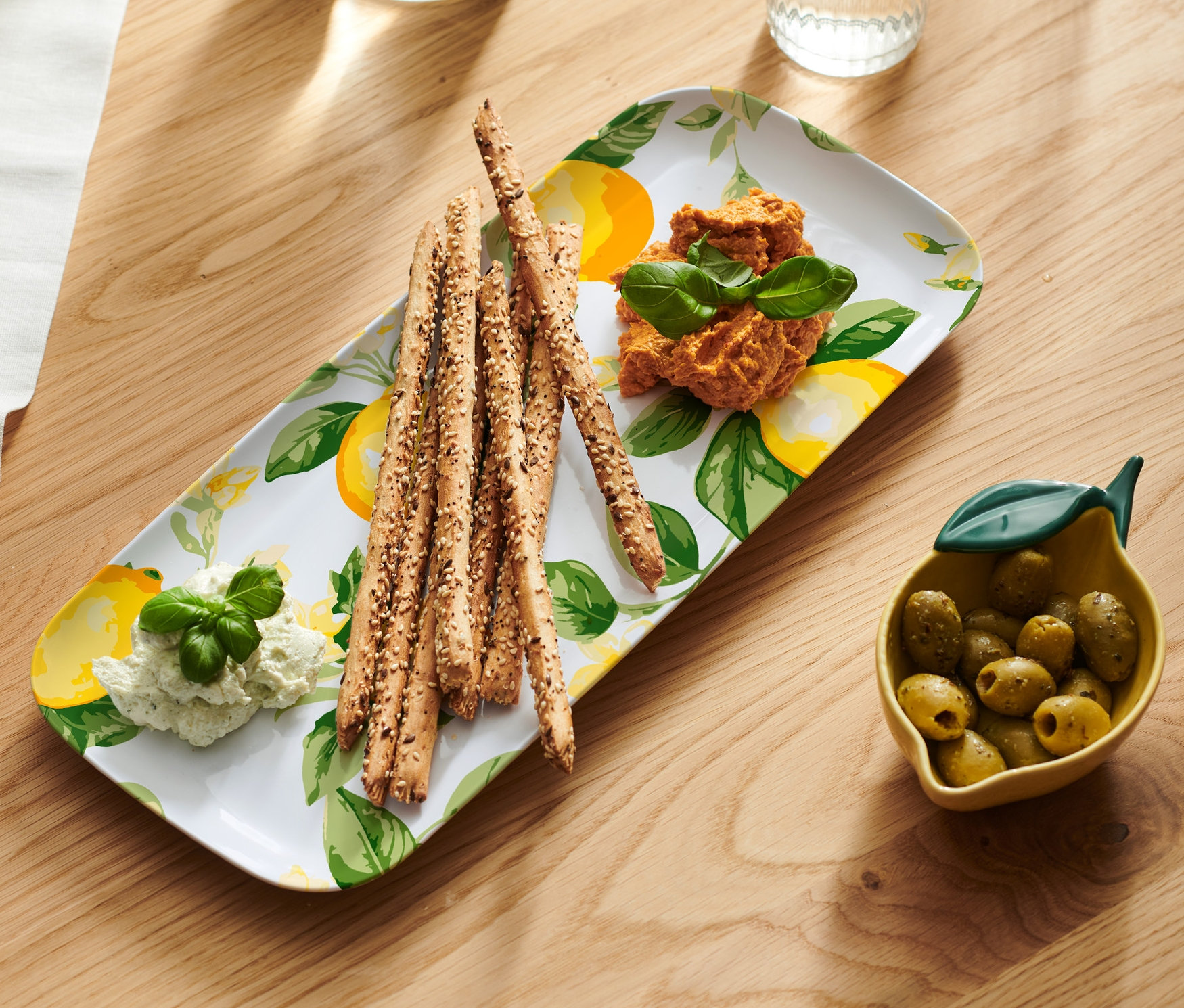 Egy fa asztalon egy kínálótálon kenyérrudak, kétféle mártás és egy tál olajbogyó látható.