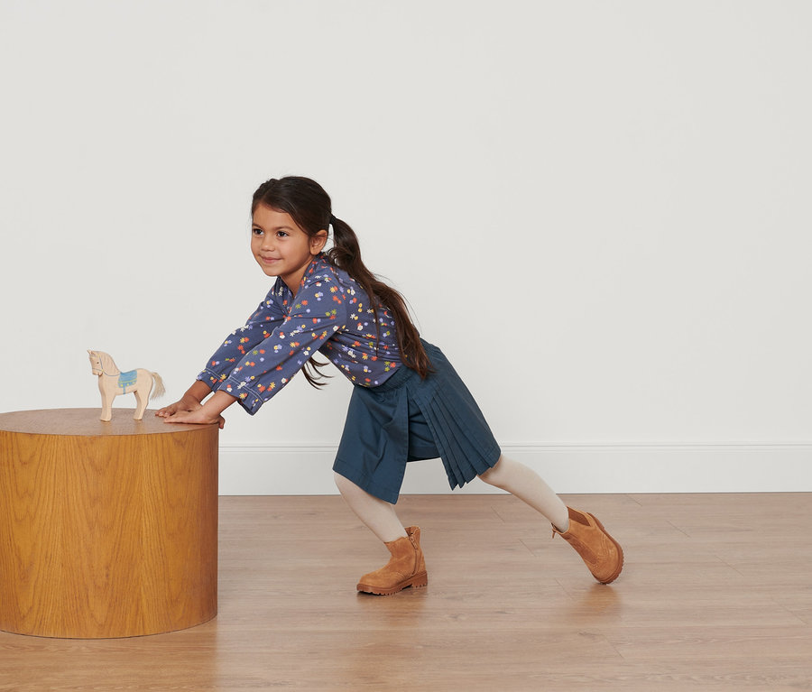 Kislány egy asztalnak dől fa lóval, kék virágos ingben és sötétkék kisgyerek skót szoknyában.