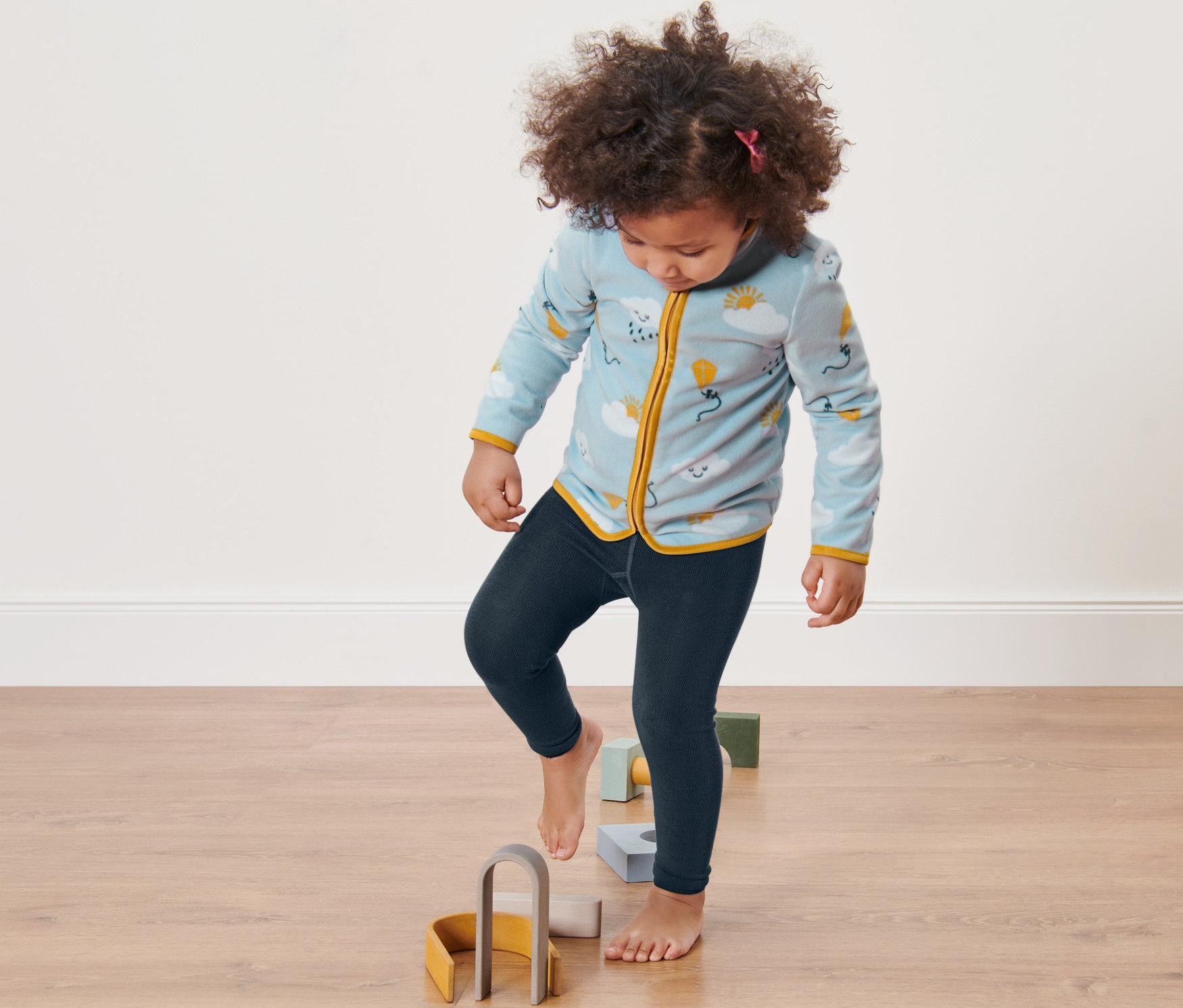 Egy lány a padlón lévő fa játékokra lép. 2 kisgyerek kötött leggings van rajta.