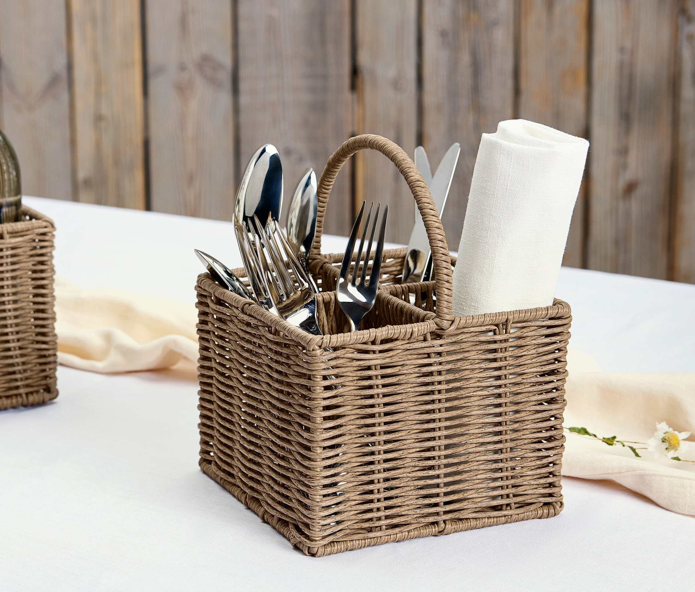 Egy fonott evőeszköztartó kosár szalvétákkal és ezüst evőeszközökkel egy fehér asztalon.