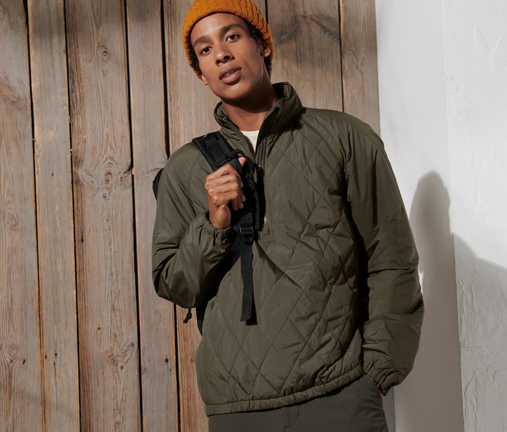 Férfi pózol egy fa fal előtt, olíva színű, félcipzáras széldzsekit és sárga sapkát visel.
