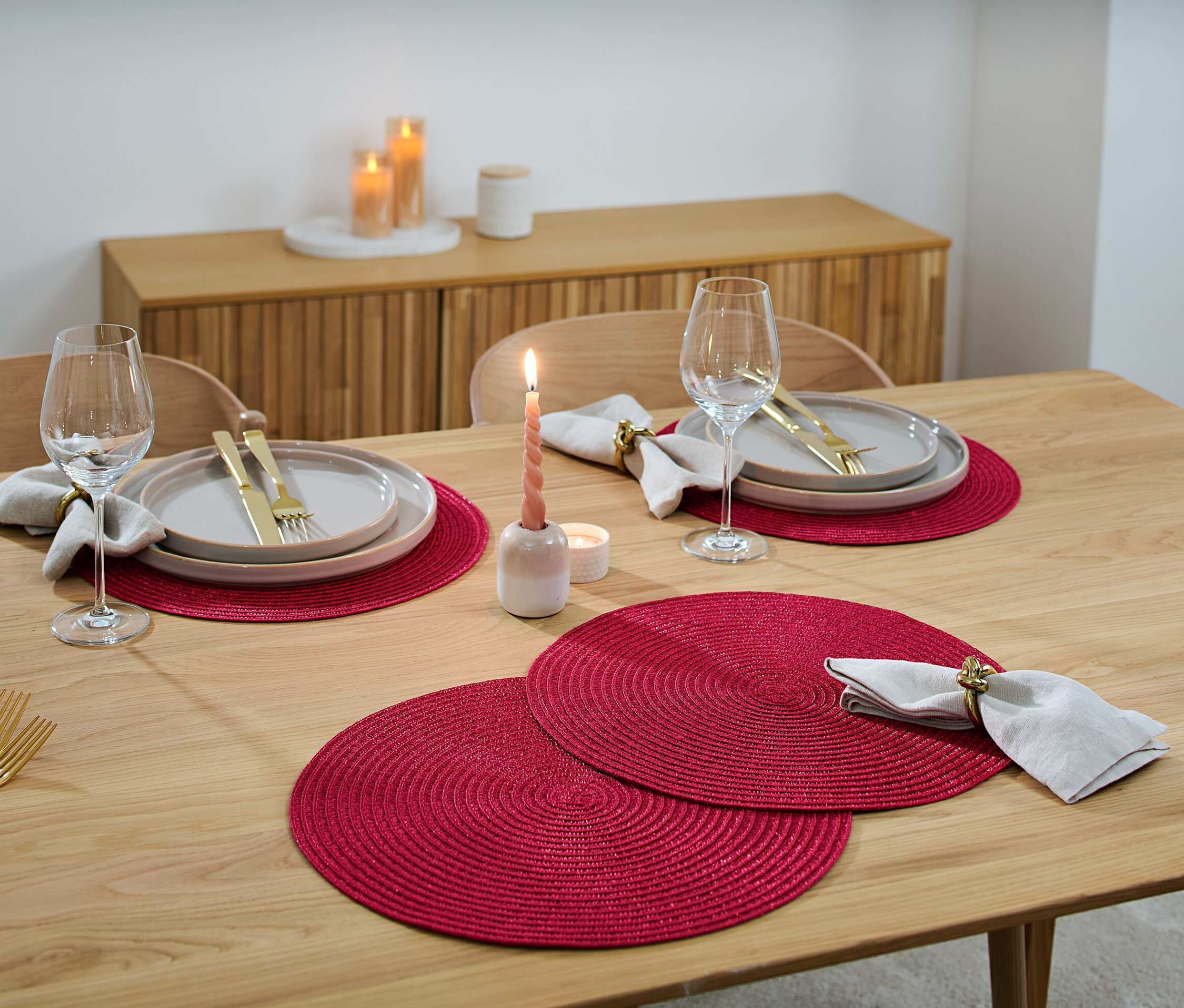 A fa asztalon terítékek találhatók 4 tányéralátéttel, tányérokkal, borospoharakkal, evőeszközökkel és gyertyákkal.
