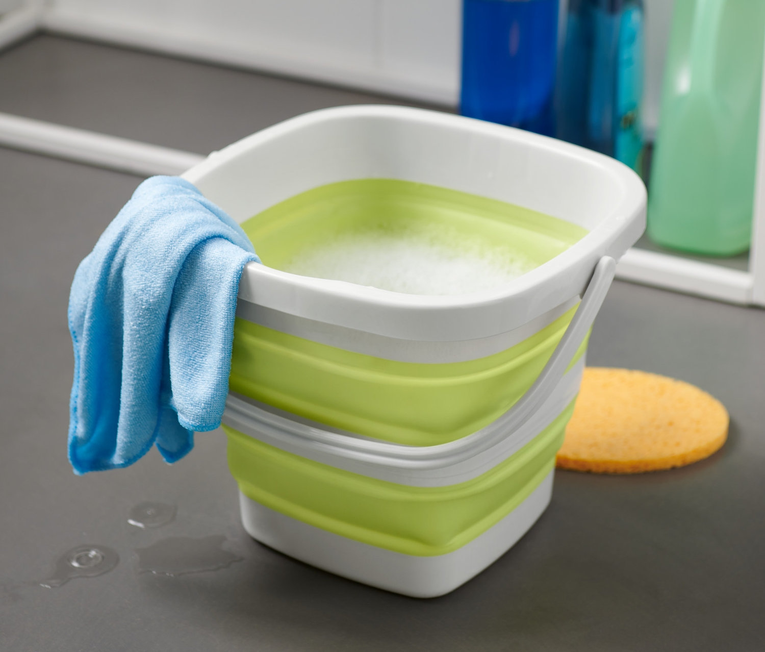 Egy szögletes, összehajtható vödör egy mosogatóronggyal és szivaccsal a padlón.