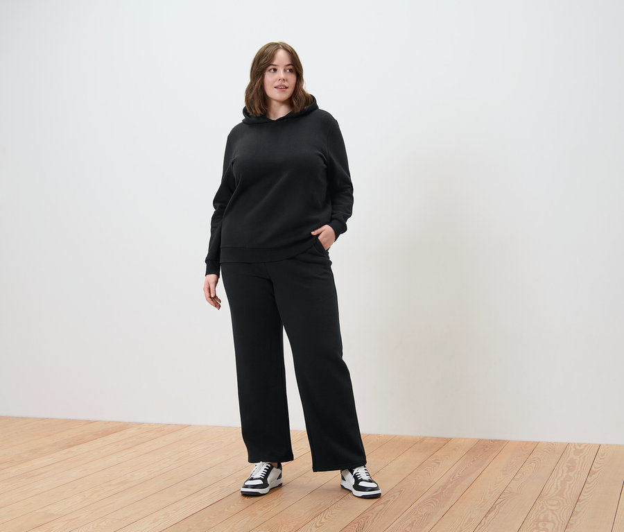 Nő fekete kapucnis szettben, bő szárú nadrággal, fa padlón állva.
