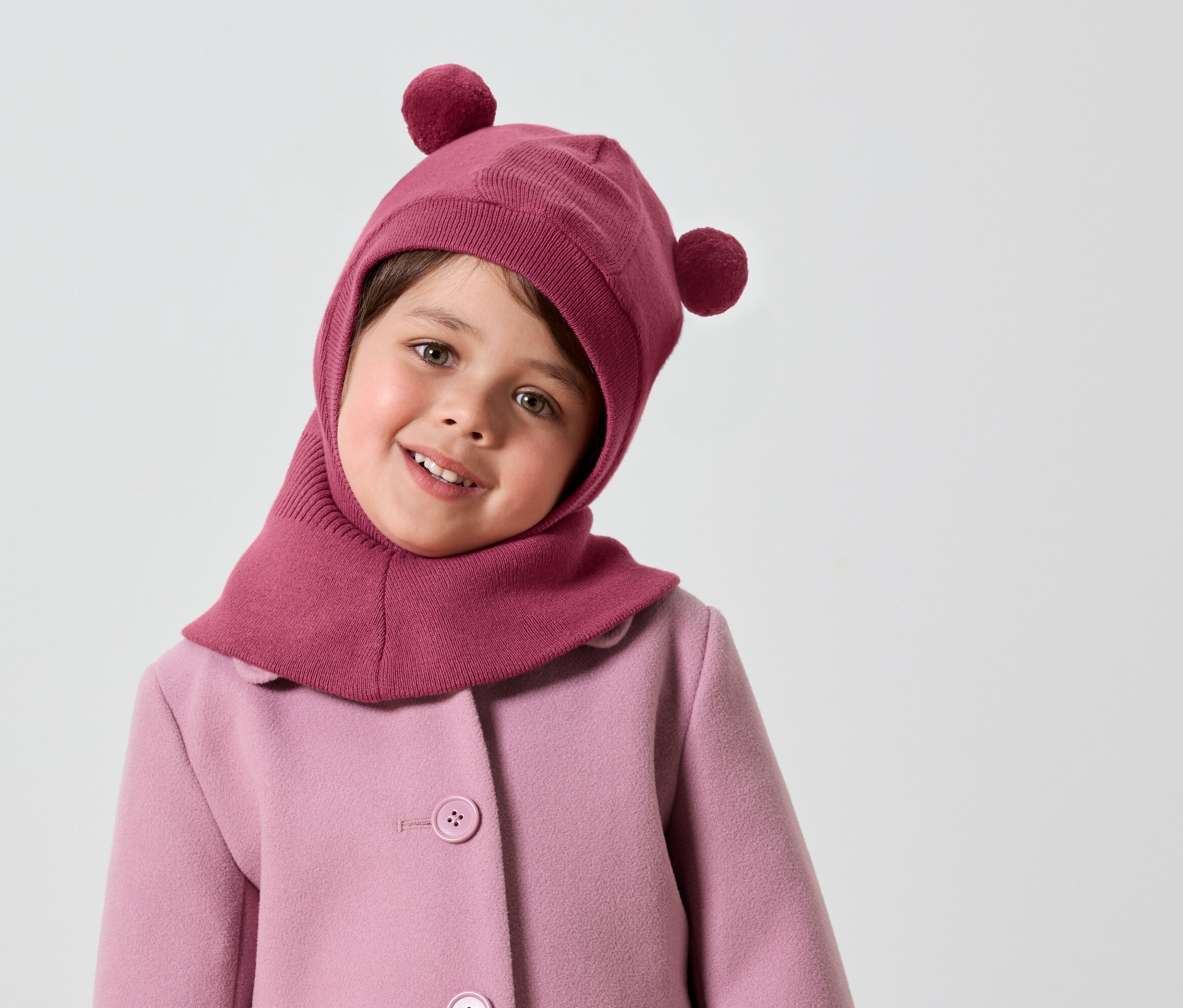 Mosolygó kislány rózsaszín, pomponos kötött sapkában és rózsaszín kabátban.