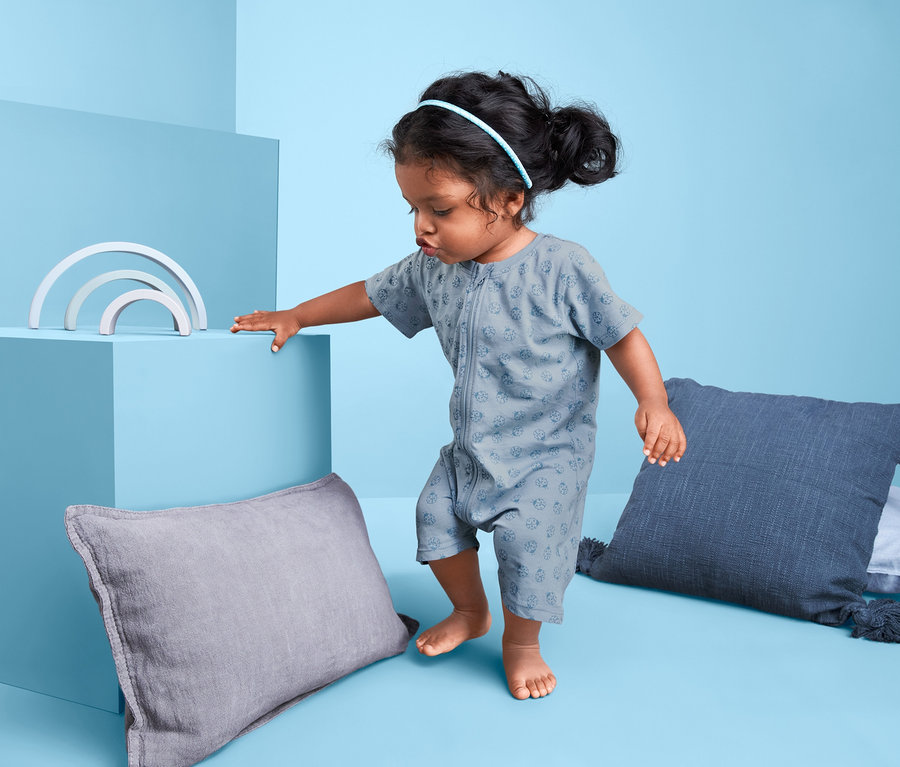 Kisgyermek zöld-kék 2 részes rövid pizsamában áll párnák és játékok mellett.