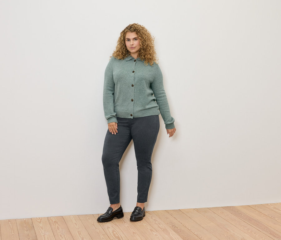  göndör hajú nő áll zöld kardigánban, szürke nadrágban és fekete mokaszinban.