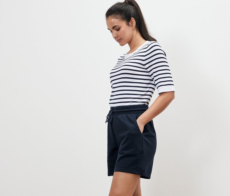 Nő csíkos pólóban és kék szabadidő rövidnadrágban áll oldalra fordulva.