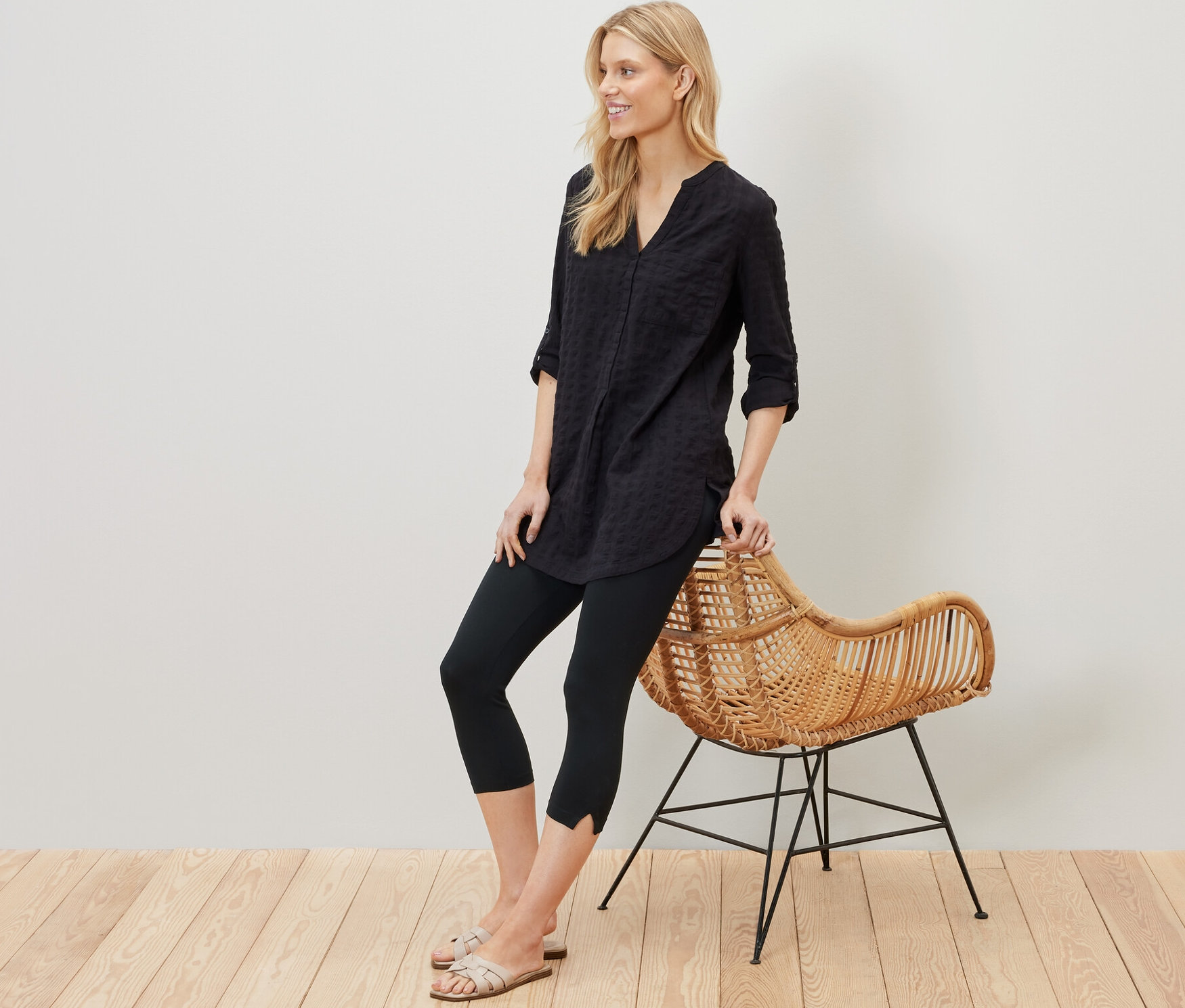 Egy szőke nő egy rattan széknek támaszkodik, és egy fekete, 3/4-es leggingset visel dekoratív elemmel.