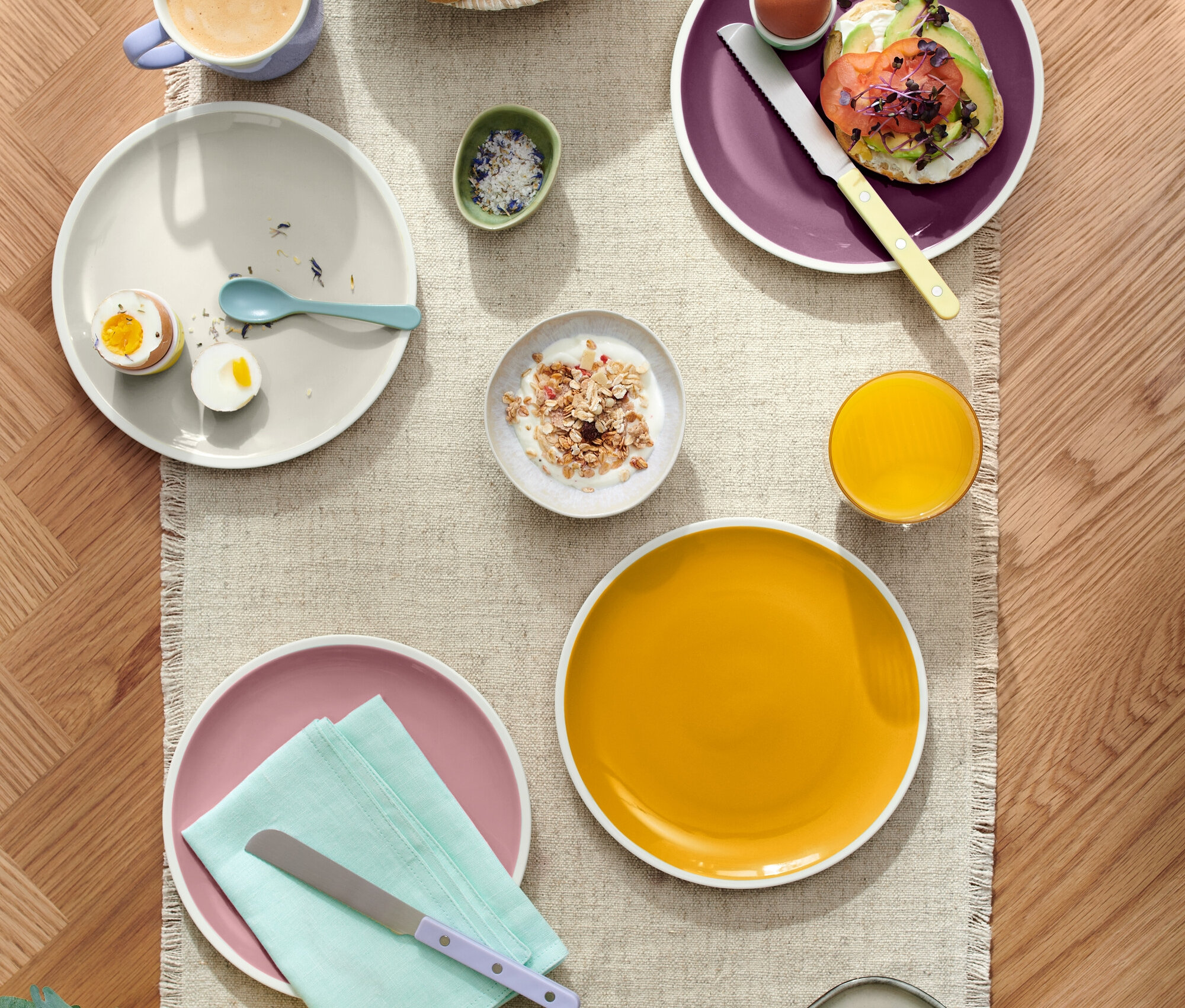 Felülnézet egy megterített asztalra, különféle ételekkel, beleértve a péksüteményeket egy kosárban, tojásos tányérokat, avokádós pirítóst és sajtot, valamint olyan italokat, mint a kávé és a gyümölcslé.