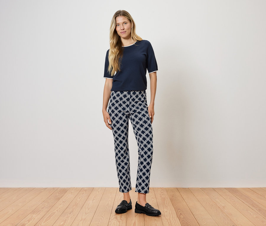 Egy nő áll egy fapadlón, sötétkék pólót és mintás nadrágot visel.