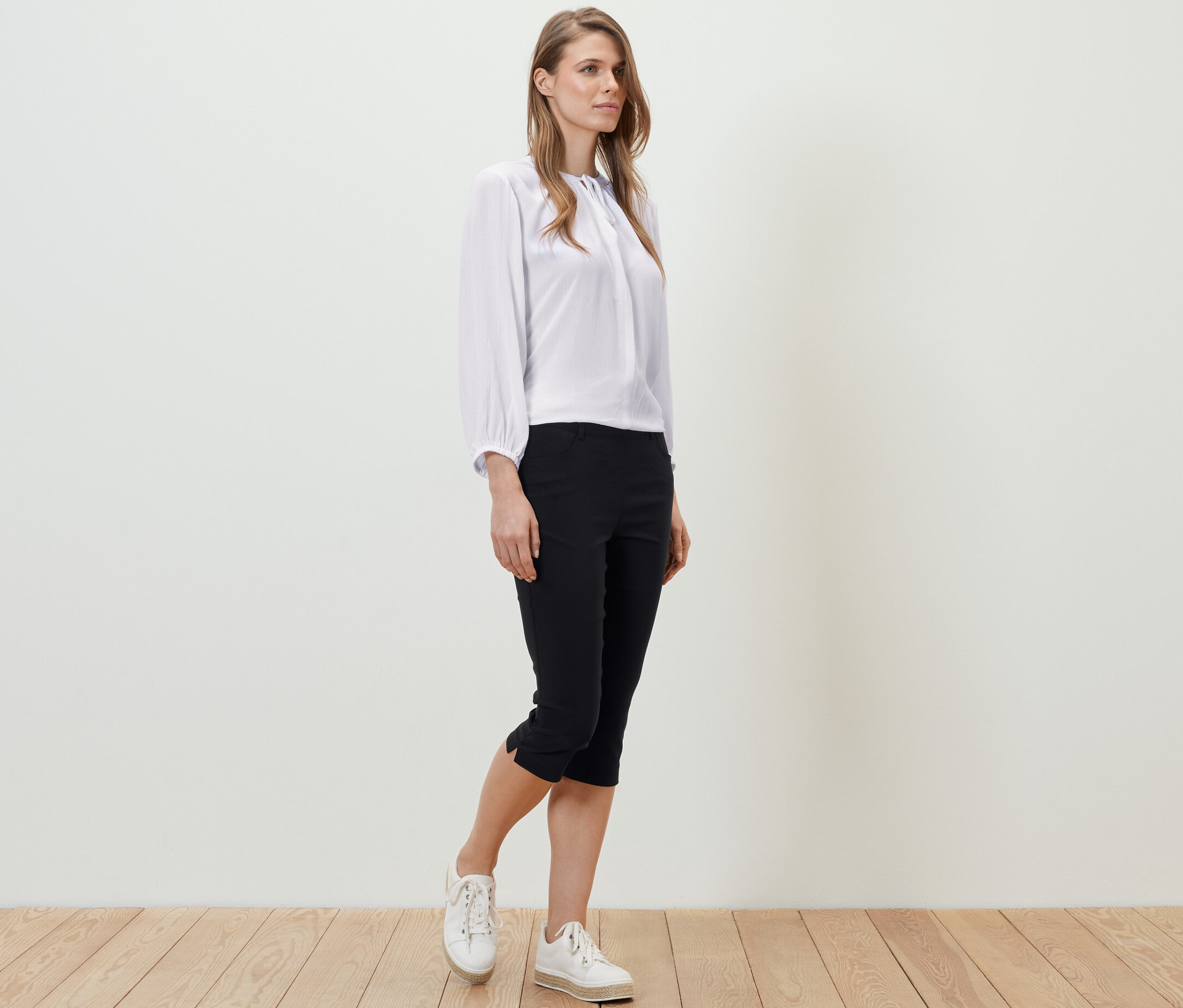 Egy nő áll a fapadlón, fehér blúzt és fekete 3/4-es bengalin nadrágot visel.