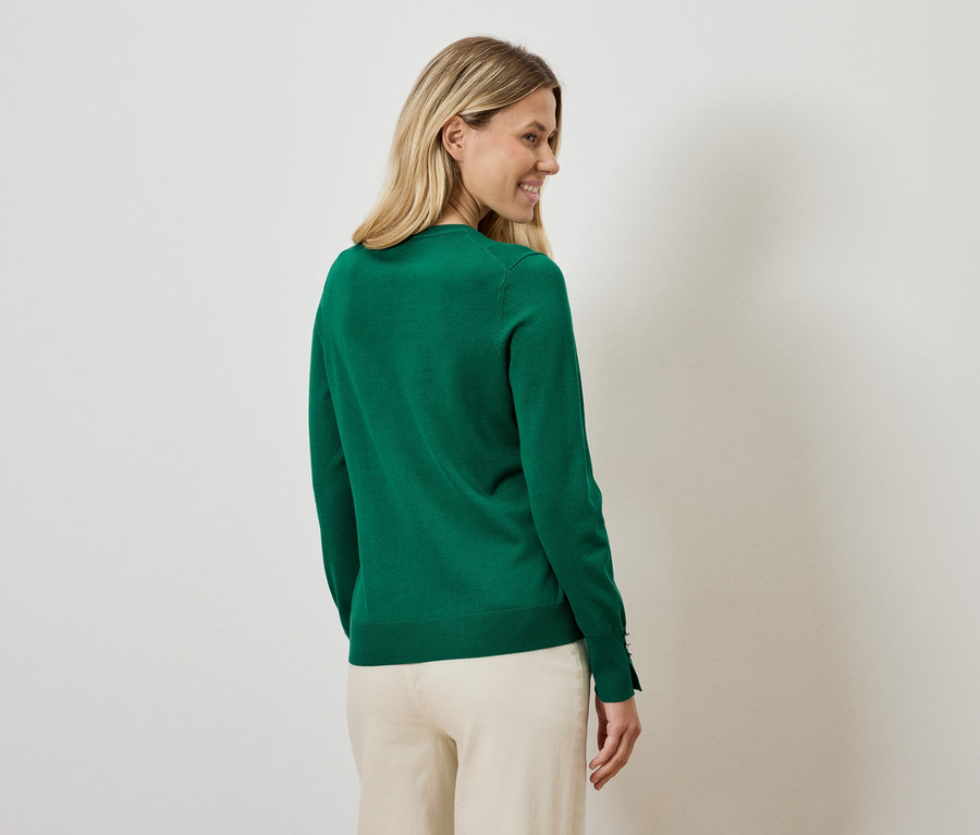 Egy nő a válla fölött néz hátra, zöld pulóvert és bézs nadrágot visel.