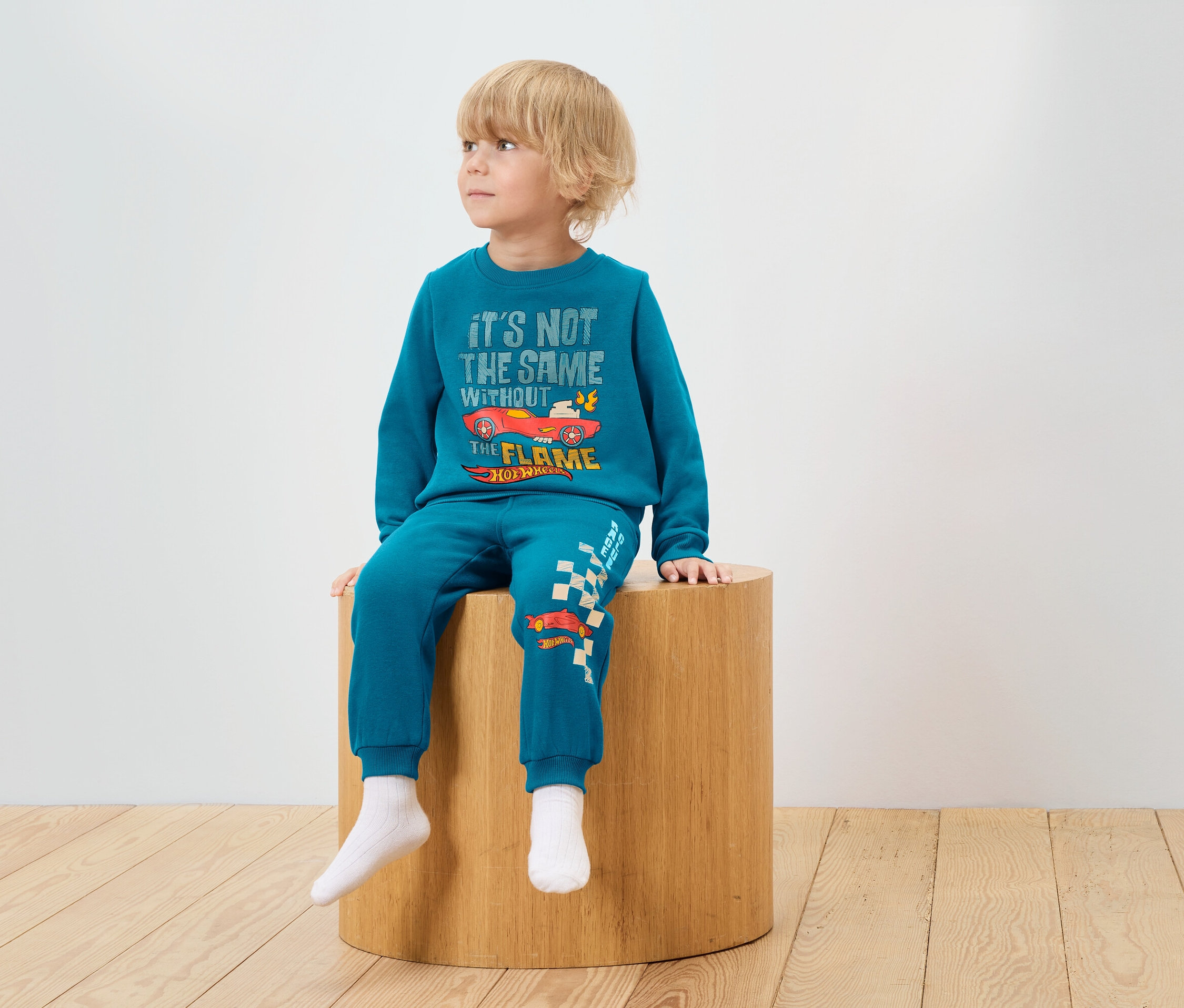 Egy fiú ül egy fa zsámolyon, és kék Hot Wheels mintás tréningruhát visel.
