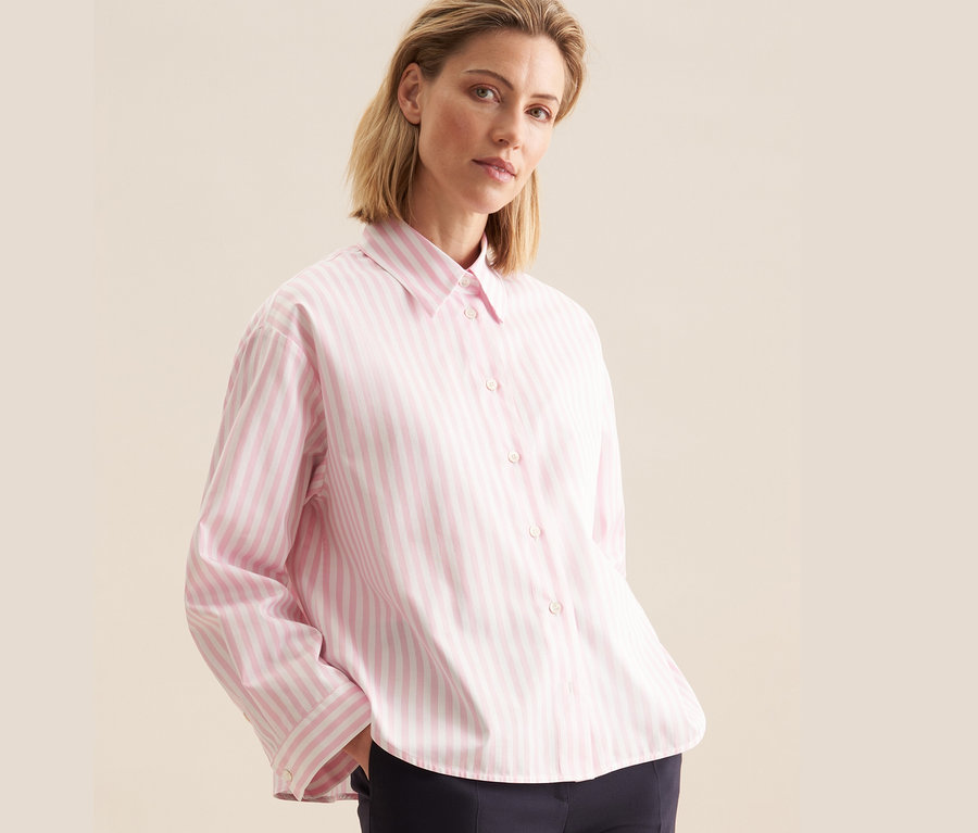 Nő visel egy rózsaszín csíkos Seidensticker túlméretes ingblúzt és egy sötétkék nadrágot.