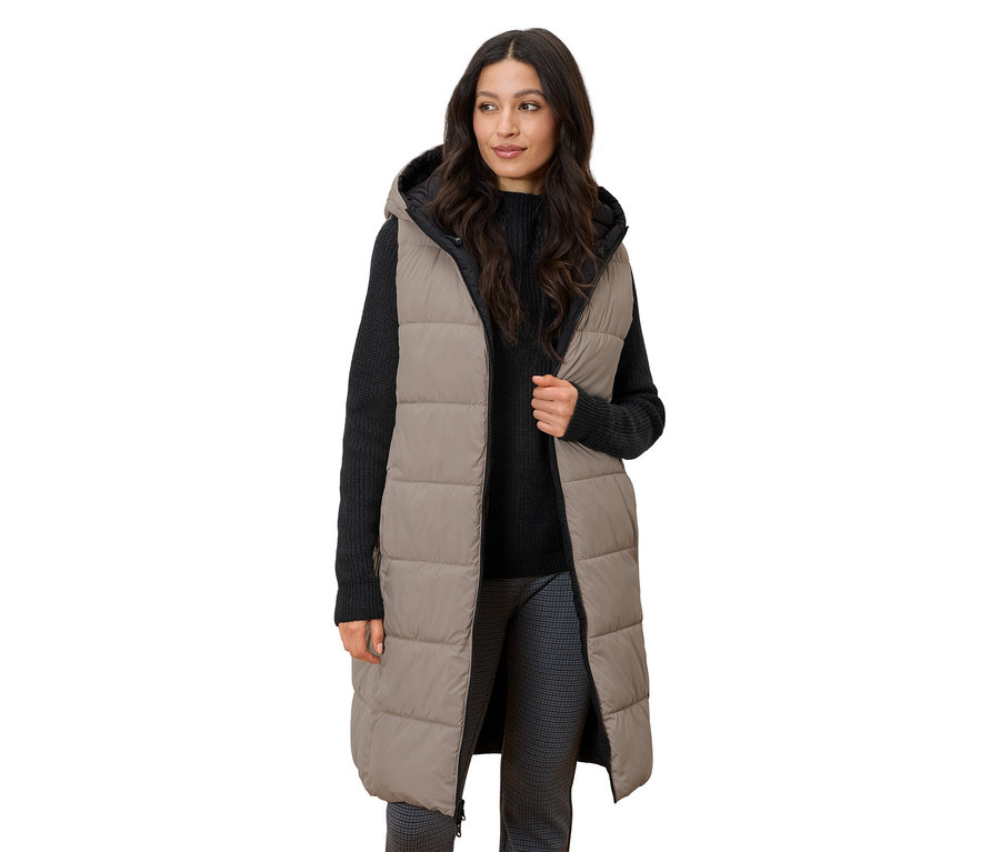 Nő áll, hosszú, steppelt, kapucnis mellényt visel.