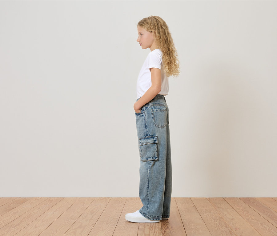 Egy szőke, göndör hajú fiatal lány áll oldalt, fehér pólót, oldalzsebes bő farmert és fehér tornacipőt visel.
