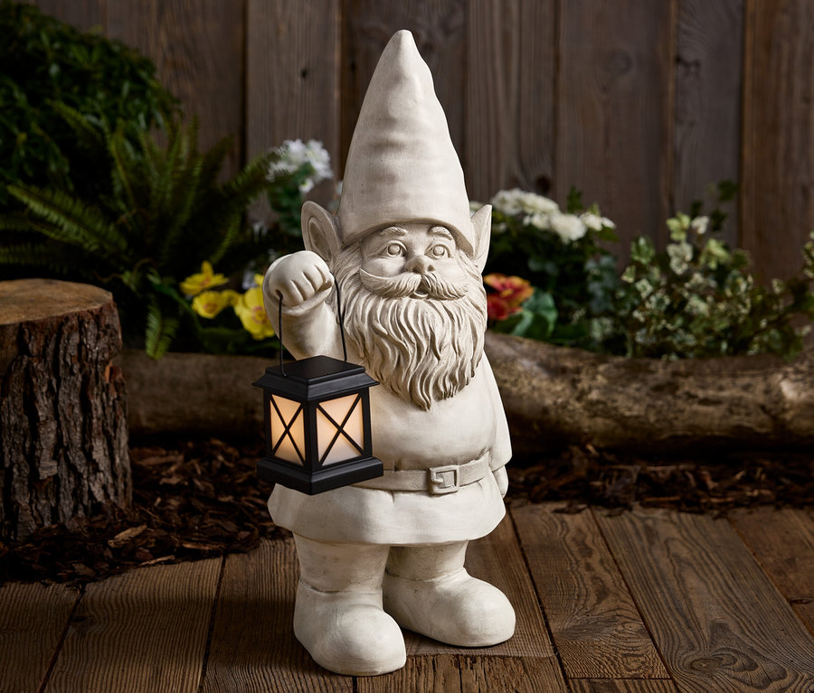 Egy kőből készült kerti törpe fekete lámpást tart egy fa háttér előtt.