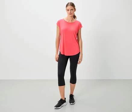 Korall színű pólót, fekete leggingset és sportcipőt viselő nő áll fehér háttér előtt.