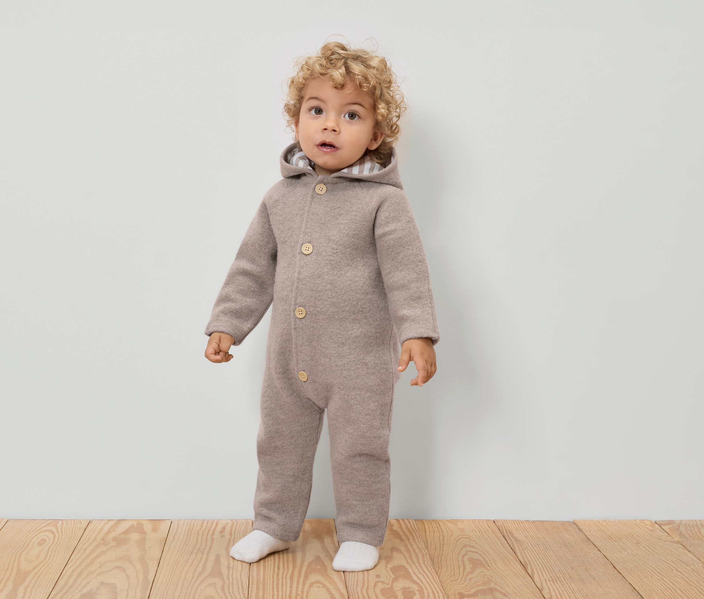 Egy göndör hajú kisgyermek világosbarna, kapucnis, gombos gyapjú overált és fehér zoknit visel.