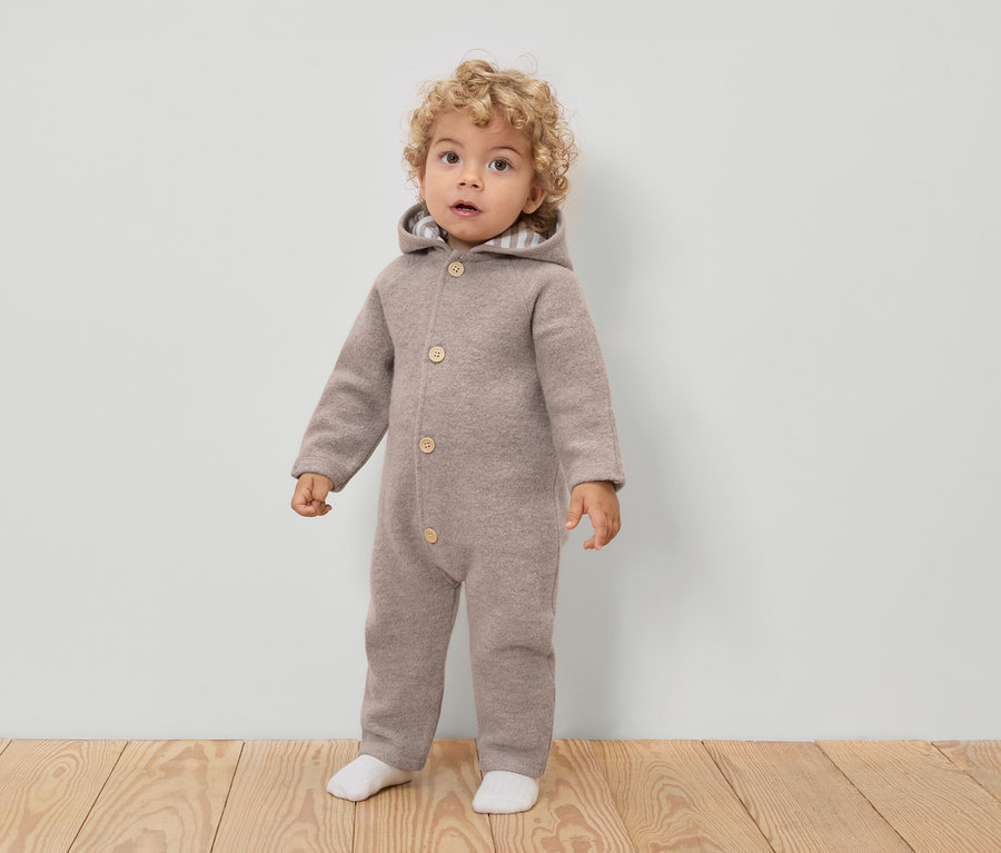 Egy göndör hajú kisgyermek világosbarna, kapucnis, gombos gyapjú overált és fehér zoknit visel.