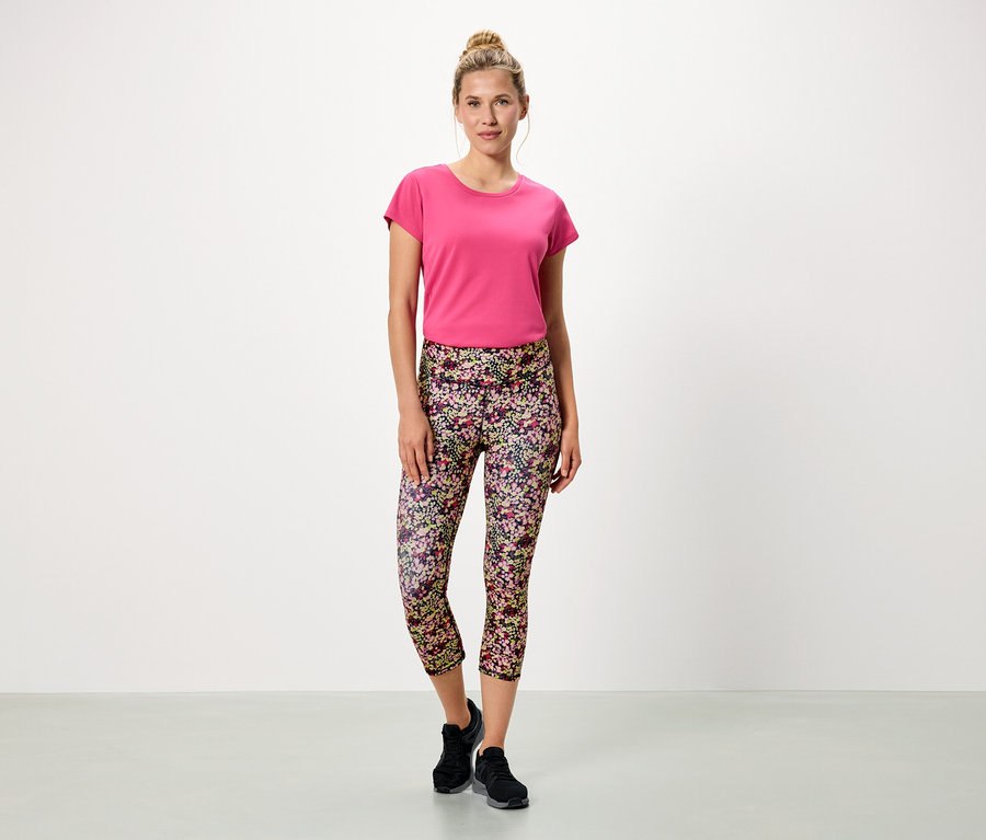 Nő áll fehér háttér előtt, rózsaszín pólót és virágos leggingst visel.