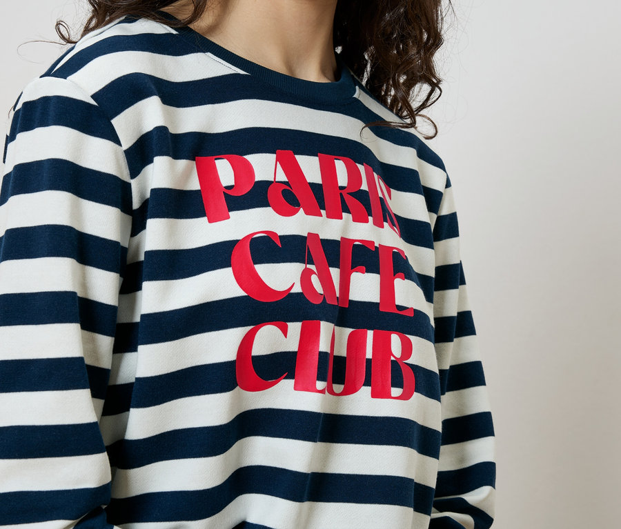 Nő visel egy kék-fehér csíkos pulóvert, rajta piros "Paris Cafe Club" felirattal.