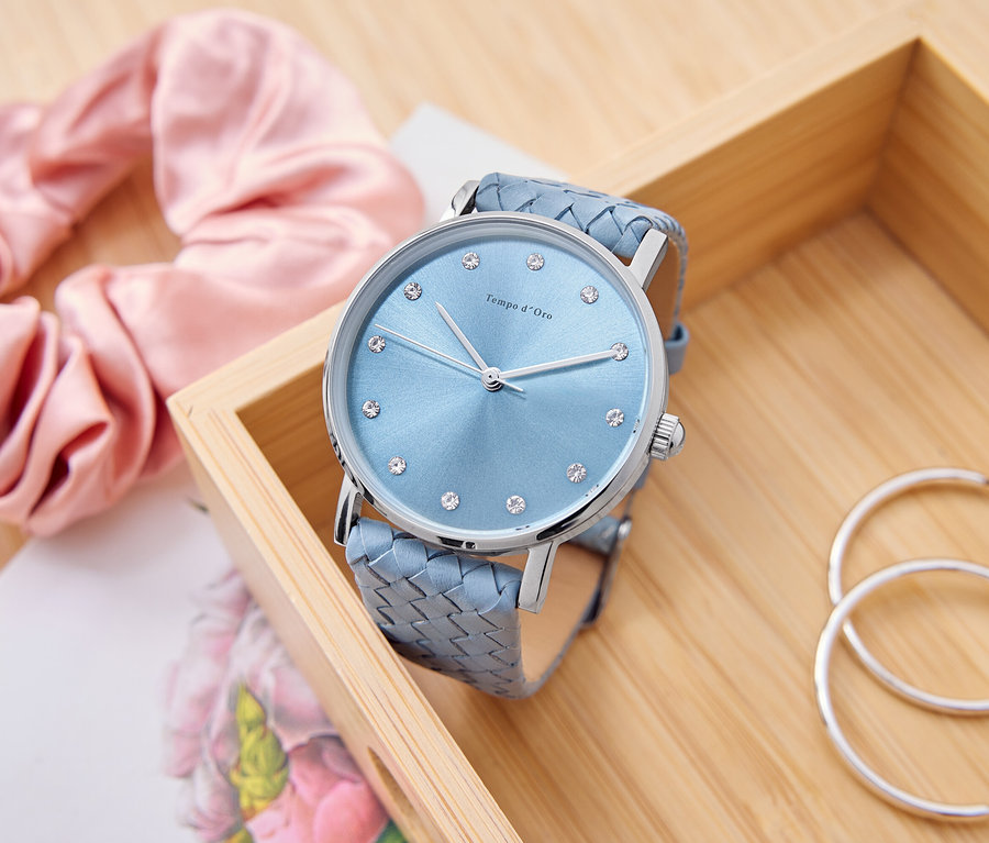 Egy világoskék, strasszköves óra fekszik egy fadobozban, mellette egy rózsaszín hajgumi, egy jegyzettömb és ezüst karika fülbevalók.