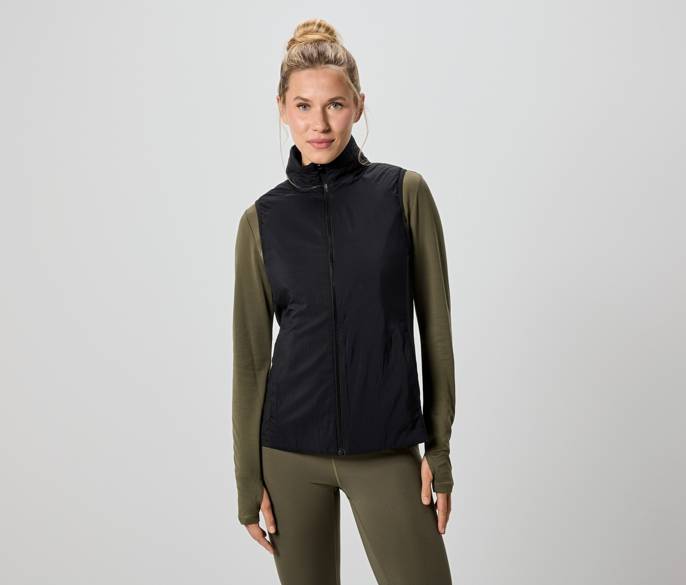 Egy szőke hajú nő fekete mellényt visel egy zöld hosszú ujjú felső és leggings felett.